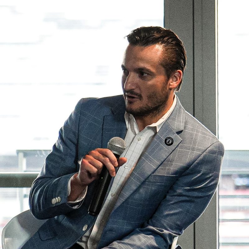 Anthony Di Fino speaking with a microphone at a conference, a Rockbeach pin on his lapel.